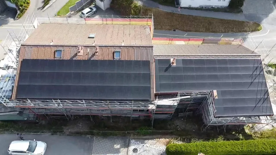 Vue aérienne : immeuble mixte à Farvagny avec modules solaires noirs sur le toit en tuiles sombres, maisons voisines autour